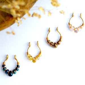 Puede incluir: Cuatro piercings de nariz dorados con detalles de cuentas. Los piercings presentan cuentas de varios colores, incluyendo azul, amarillo y rosa. Las joyas se exhiben sobre una superficie blanca, con un fondo borroso.