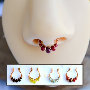 Puede incluir: Un anillo de nariz de color cobre con cuentas rojas se muestra en un maniquí. Debajo, se muestran tres anillos de nariz adicionales, con cuentas negras, amarillas y turquesas. La joyería está diseñada con un estilo de alambre envuelto.