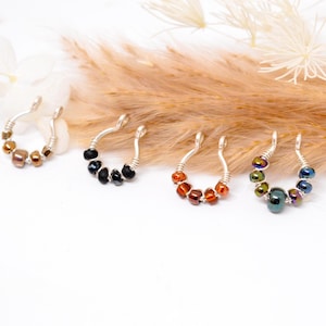 Puede incluir: Cuatro piercings de nariz plateados, cada uno adornado con cuentas de diferentes colores. Las cuentas incluyen tonos de marrón, negro, naranja y colores iridiscentes. Las joyas se exhiben sobre una superficie blanca con acentos florales secos.