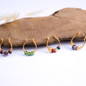 Puede incluir: Cuatro anillos de nariz dorados con cuentas de diferentes colores. Los anillos de nariz están dispuestos en fila, cada uno con una combinación única de cuentas moradas, verdes, rojas y azules. Las joyas se exhiben sobre una superficie de madera.