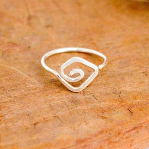 May include: A silver ring with a square spiral design. The ring is set on a wooden surface with dried, fluffy, cream-colored plants. The ring's design features a square shape with a spiral in the center. The wooden surface has a natural wood grain.