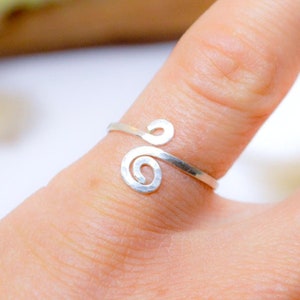 Può includere: Un anello color argento con un design a spirale è indossato su un dito. L'anello è delicato e sembra essere fatto di filo. L'anello è il punto focale dell'immagine, con la mano e lo sfondo leggermente sfocati.