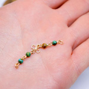 May include: A delicate gold-colored jewelry piece featuring a decorative floral design and small, round beads in shades of teal and brown. The beads are strung along a curved chain, creating an elegant and intricate accessory. The piece is held in a hand.