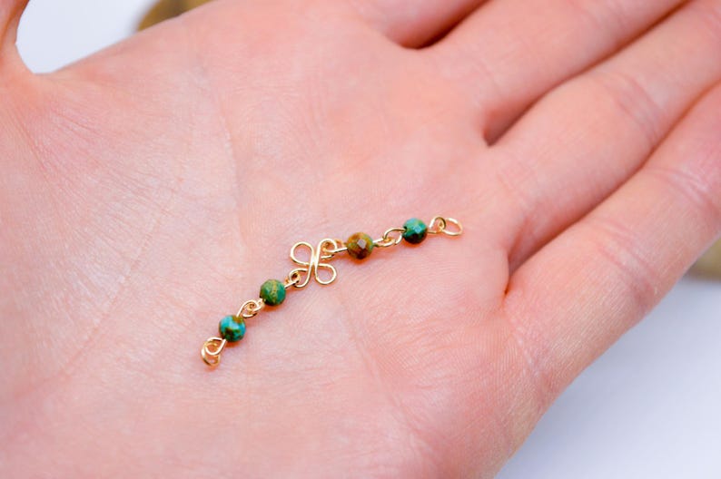 May include: A delicate gold-colored jewelry piece featuring a decorative floral design and small, round beads in shades of teal and brown. The beads are strung along a curved chain, creating an elegant and intricate accessory. The piece is held in a hand.