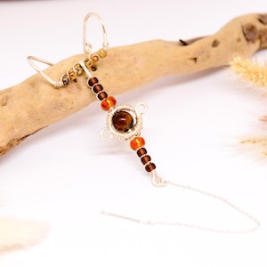 May include: A silver-toned ear cuff with a dangling chain, adorned with amber and brown beads. The design features a central circular element with a large, round, amber-colored stone. The cuff is placed on a piece of driftwood.