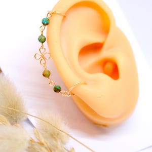 May include: A gold-colored ear cuff with a delicate chain and turquoise-colored beads. The cuff is attached to a small hoop earring and a decorative floral charm. The jewelry is displayed on a mannequin ear.