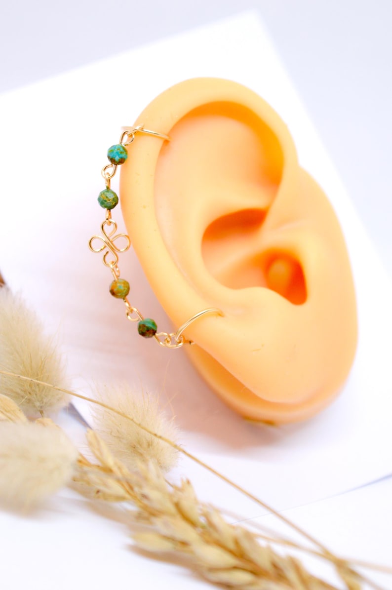 May include: A gold-colored ear cuff with a delicate chain and turquoise-colored beads. The cuff is attached to a small hoop earring and a decorative floral charm. The jewelry is displayed on a mannequin ear.