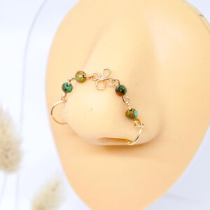 May include: A gold-colored nose ring with a delicate design. The nose ring features a series of small, round, green and brown beads connected by a thin wire. The design includes a decorative swirl and a ring that fits around the nostril.