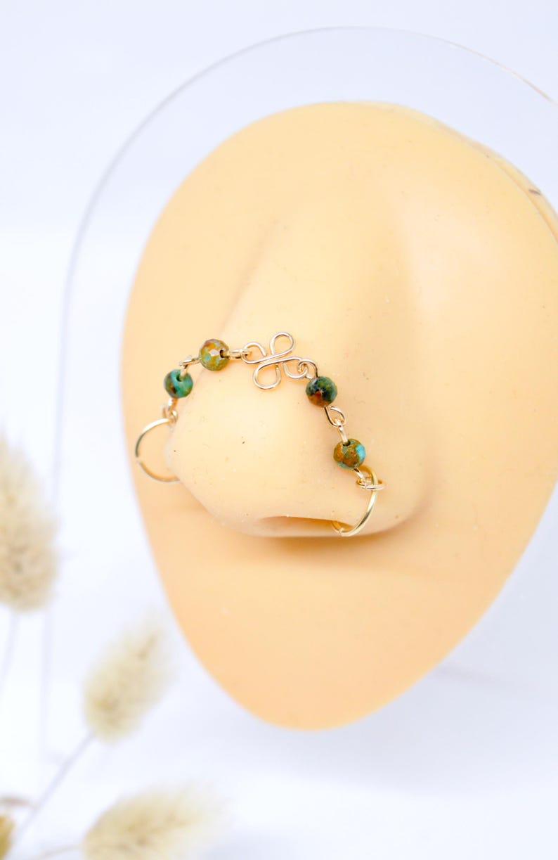 May include: A gold-colored nose ring with a delicate design. The nose ring features a series of small, round, green and brown beads connected by a thin wire. The design includes a decorative swirl and a ring that fits around the nostril.