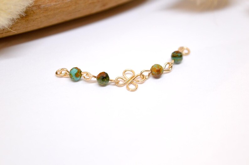 May include: A gold-colored jewelry component with a decorative floral design and small, round, turquoise and brown beads. The beads are spaced along a chain-like structure.