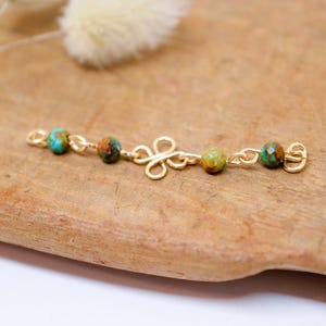 May include: A gold-colored jewelry piece with turquoise and brown beads. The piece has a floral-shaped center and is displayed on a wooden surface. The beads are spaced along the gold chain.