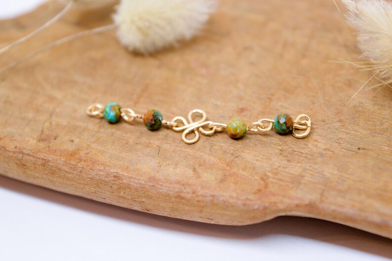 May include: A gold-colored jewelry piece with turquoise and brown beads. The piece has a floral-shaped center and is displayed on a wooden surface. The beads are spaced along the gold chain.