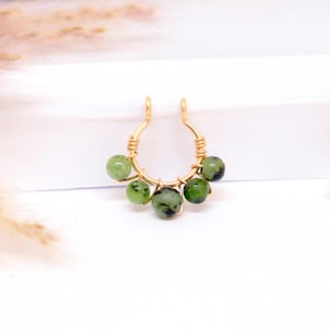 Puede incluir: Un aro de nariz dorado, envuelto en alambre, con cinco cuentas de piedras preciosas verdes. Las cuentas, de diferentes tamaños y tonos de verde, añaden un toque natural y único a la joya.