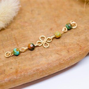 May include: A gold-colored jewelry piece with a clover-shaped design and small, colorful beads. The beads are a mix of turquoise, brown, and green. The piece is displayed on a wooden surface.