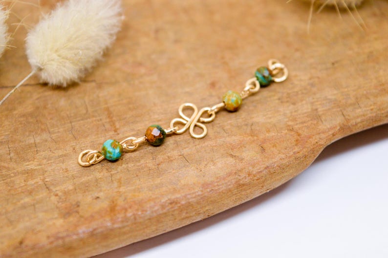 May include: A gold-colored jewelry piece with a clover-shaped design and small, colorful beads. The beads are a mix of turquoise, brown, and green. The piece is displayed on a wooden surface.