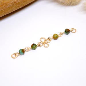 May include: Gold-toned jewelry component with a decorative floral-like center and chain links. The chain holds five round, multi-colored beads in shades of green, brown, and turquoise. The piece is set against a white background.
