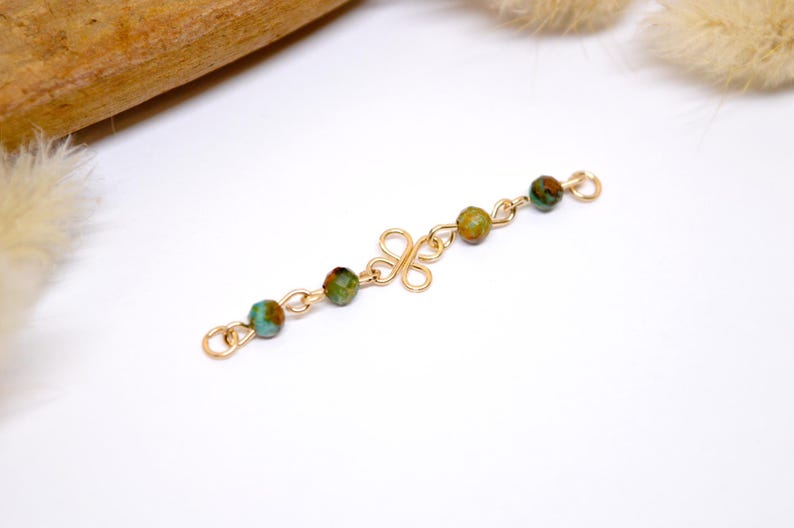 May include: Gold-toned jewelry component with a decorative floral-like center and chain links. The chain holds five round, multi-colored beads in shades of green, brown, and turquoise. The piece is set against a white background.