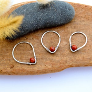 Könnte beinhalten: Drei silberne Creolen mit einer kleinen, runden, rotbraunen Perle. Die Ohrringe werden auf einer Holzoberfläche mit einem dunkelgrauen Stein und getrockneten Pflanzen im Hintergrund präsentiert.