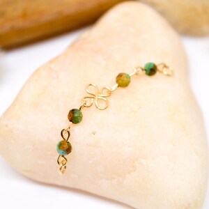 May include: A delicate gold-colored bracelet featuring a four-leaf clover design and small, round beads in shades of green and brown. The bracelet is displayed on a light-colored, smooth stone, showcasing its intricate details.
