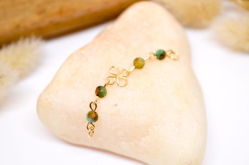 May include: A delicate gold-colored bracelet featuring a four-leaf clover design and small, round beads in shades of green and brown. The bracelet is displayed on a light-colored, smooth stone, showcasing its intricate details.