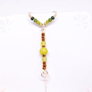 May include: A handcrafted silver-toned ring with a dangling charm. The ring features a series of green and yellow beads, with copper-colored accents. The beads are arranged in a vertical line, creating a unique and eye-catching design.