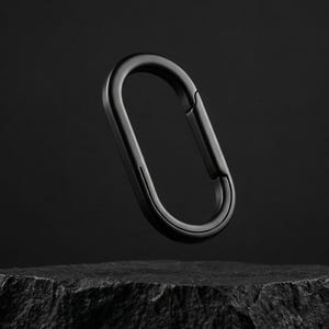 Puede incluir: Un mosquetón negro con acabado mate, suspendido en el aire sobre un fondo oscuro. El clip de forma ovalada es de metal y tiene una puerta con resorte para un cierre seguro. El clip está apoyado sobre una roca negra.