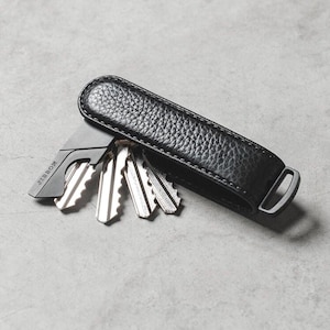Op de afbeelding: Een zwarte leren sleutelorganizer met een zilveren metalen sluiting en meerdere sleutels. De organizer heeft een getextureerd oppervlak en een minimalistisch ontwerp, perfect om sleutels veilig en georganiseerd te houden.