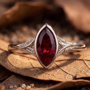 May include: A silver-toned ring featuring a central marquise-cut deep red gemstone set in a bezel. The band displays an intricate, engraved leaf-like pattern extending from the setting along both sides of the metal shank.