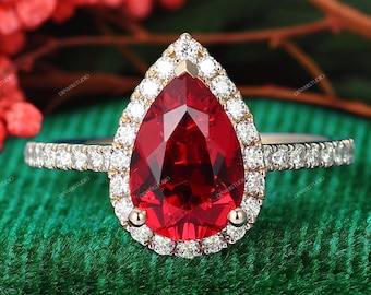 May include: A pear-shaped red gemstone ring set in a gold band. The central red stone is surrounded by a halo of small, round, clear white diamonds. The band is also encrusted with a row of small, round, clear white diamonds along the sides.