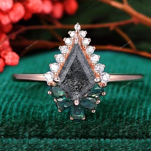May include: A rose gold ring featuring a large, kite-shaped, dark gray gemstone, surrounded by small white diamonds and green gemstones. The ring is set against a green velvet background, with red berries in the background.