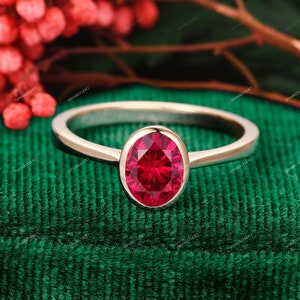 May include: A delicate gold ring with a bezel setting featuring a vibrant red oval gemstone. The ring is displayed on a green velvet surface.