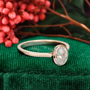 Op de afbeelding: Een verlovingsring van ros&eacute;goud met een ovale diamant in een bezel-setting. De ring ligt op een groene fluwelen ondergrond.