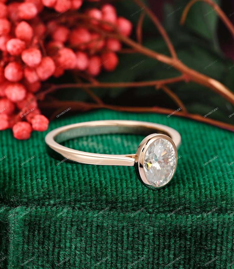 Op de afbeelding: Een verlovingsring van ros&eacute;goud met een ovale diamant in een bezel-setting. De ring ligt op een groene fluwelen ondergrond.