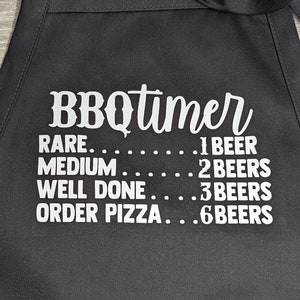 May include: Black BBQ apron with white text that reads "BBQtimer" and a list of doneness levels for meat with corresponding beer counts: "RARE...1 BEER", "MEDIUM......2 BEERS", "WELL DONE....3 BEERS", "ORDER PIZZA...6 BEERS".