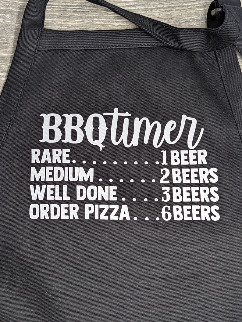 May include: Black BBQ apron with white text that reads "BBQtimer" and a list of doneness levels for meat with corresponding beer counts: "RARE...1 BEER", "MEDIUM......2 BEERS", "WELL DONE....3 BEERS", "ORDER PIZZA...6 BEERS".
