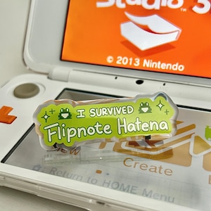 May include: A clear sticker with the text "I SURVIVED Flipnote Hatena" in green and white, featuring pixelated frog graphics. The sticker is placed on a white Nintendo 3DS console, with the Flipnote Studio 3D game visible on the screen.