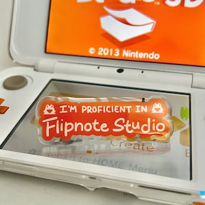 May include: A white Nintendo 3DS handheld game console displaying the Flipnote Studio 3D logo on the top screen. A clear sticker with the text "I'M PROFICIENT IN Flipnote Studio" is on the bottom screen. The sticker is orange and white.
