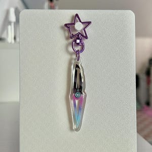 May include: A clear acrylic keychain featuring a dagger design with a purple, blue, and pink gradient. The keychain has a purple star-shaped clasp and a circular attachment. The dagger has a black and silver design.