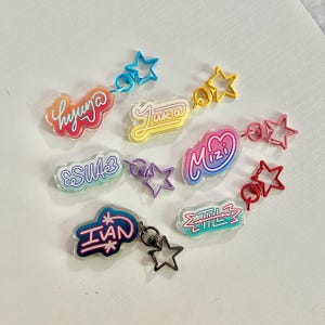 May include: A collection of colorful acrylic keychains with various names and star-shaped accents. Each keychain features a different name in a unique font and color scheme, connected to a star charm via a small ring.