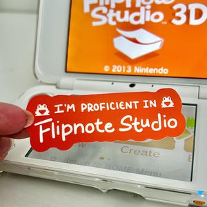 May include: An orange sticker with the text "I'M PROFICIENT IN Flipnote Studio" is held over a Nintendo 3DS. The 3DS screen displays "Flipnote Studio 3D" and the Nintendo logo. The sticker has a pixelated frog and a pixelated game controller.