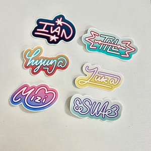 May include: A collection of six colorful stickers with various names in stylized fonts. The stickers feature a range of colors, including blue, red, yellow, and pink, with white outlines. The stickers are on a white background.