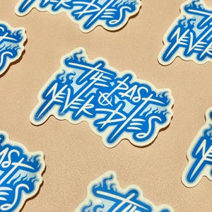 May include: Blue and white sticker with the text "The Past Never Dies" in a stylized font with flames around the text.