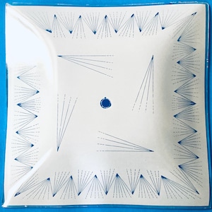 May include: A square glass plate featuring a white surface with a blue geometric pattern. The border consists of repeating blue fan-like lines, while the center displays four sets of radiating lines and a small solid blue circle in the middle.