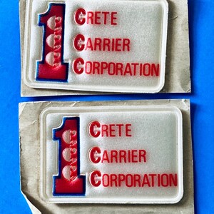 May include: Two rectangular patches featuring a large red number one with a blue outline and three circular cutouts. To the right, the red text reads Crete Carrier Corporation, with the letter C highlighted in each word. The patches are on paper.