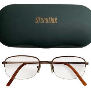 May include: A pair of brown metal-framed eyeglasses with clear lenses, resting below a dark green eyeglass case. The case has the brand name "Sferoflex" printed in gold. The glasses have a half-rim design.