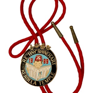 May include: A red bolo tie featuring a circular emblem with the text "R.E. Tiny Rhinehart 1999 Kerbela Temple." The emblem has a black border, gold accents, and a light blue center with a cartoon figure reading a newspaper.