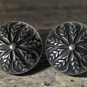 May include: A pair of round, dark silver-toned metal buttons featuring a raised, embossed wheat stalk pattern radiating from a central point. The buttons have a polished finish on the raised areas and a darkened, recessed background texture.