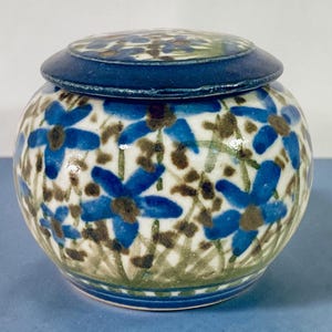 Könnte beinhalten: Ein runder Keramiktopf mit Deckel, verziert mit einem handgemalten Blumenmuster. Der Topf ist weiß mit blauen Blumen und braunen Akzenten. Der Deckel und der Rand sind einfarbig dunkelblau.