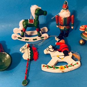 May include: A collection of Christmas ornaments on a blue background. The ornaments include a green rocking horse, a Santa Claus figure, a drummer boy, and a toy soldier on a rocking horse. Other ornaments include a red horn and a white horse head.