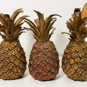 May include: Three decorative pineapple figurines arranged in a row. The two outer pineapples are finished in a metallic gold tone, while the center pineapple features a reddish-brown hue. Each piece has a textured, cross-hatched body and leaves.